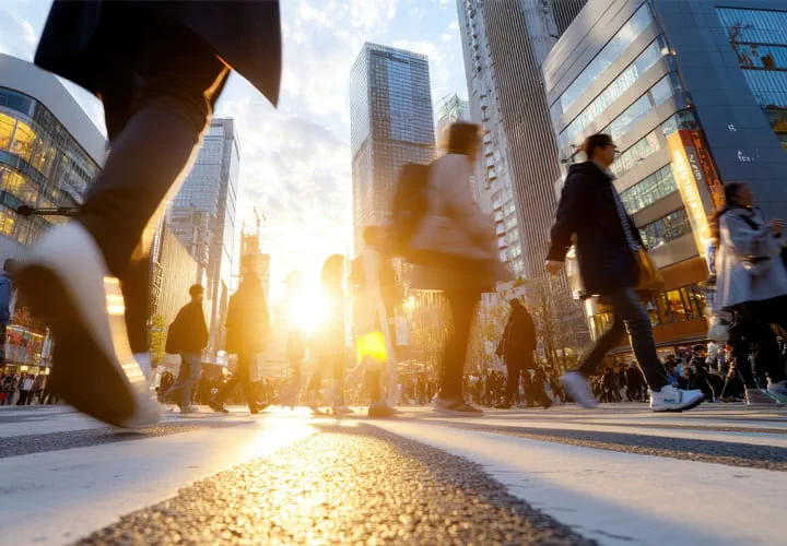 Menschen, die eine Stadtstraße mit hohen Gebäuden und Sonnenlicht im Hintergrund überqueren