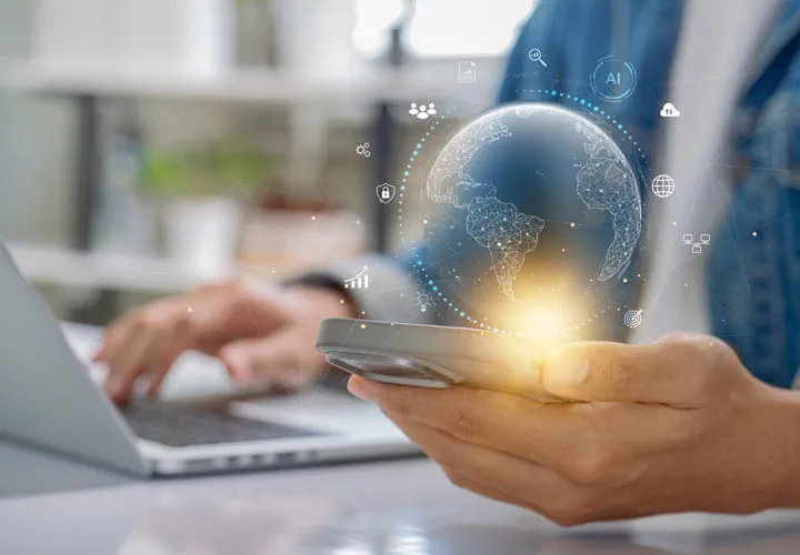 Persona sosteniendo un smartphone con un globo terráqueo digital luminoso sobre él, sentada en un escritorio con un ordenador portátil al fondo