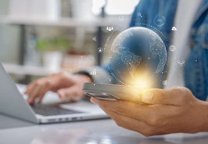 Persona sosteniendo un smartphone con un globo terráqueo digital luminoso sobre él, sentada en un escritorio con un ordenador portátil al fondo
