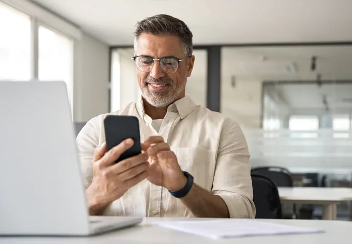 Person sitting at a desk using a smartphone, with an open laptop in front, in a modern office setting